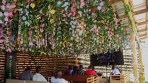 An outdoor seating area with wooden benches and tables, surrounded by greenery and hanging decorations. - Riverview Hotel and Suites (Ife)