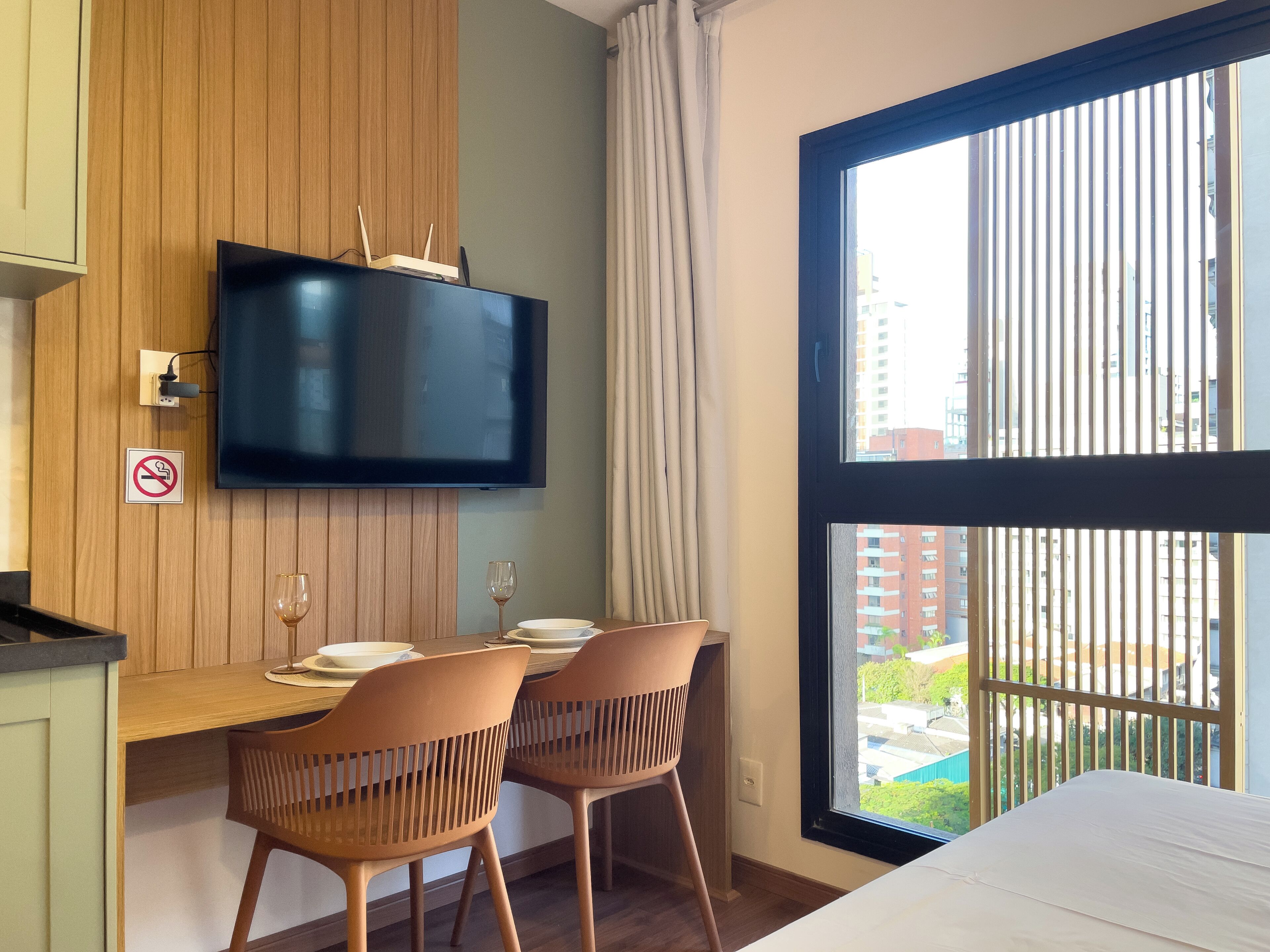 A hotel room with a TV mounted on a wooden panel wall, a dining table with chairs, and a large window with a view of buildings and greenery.