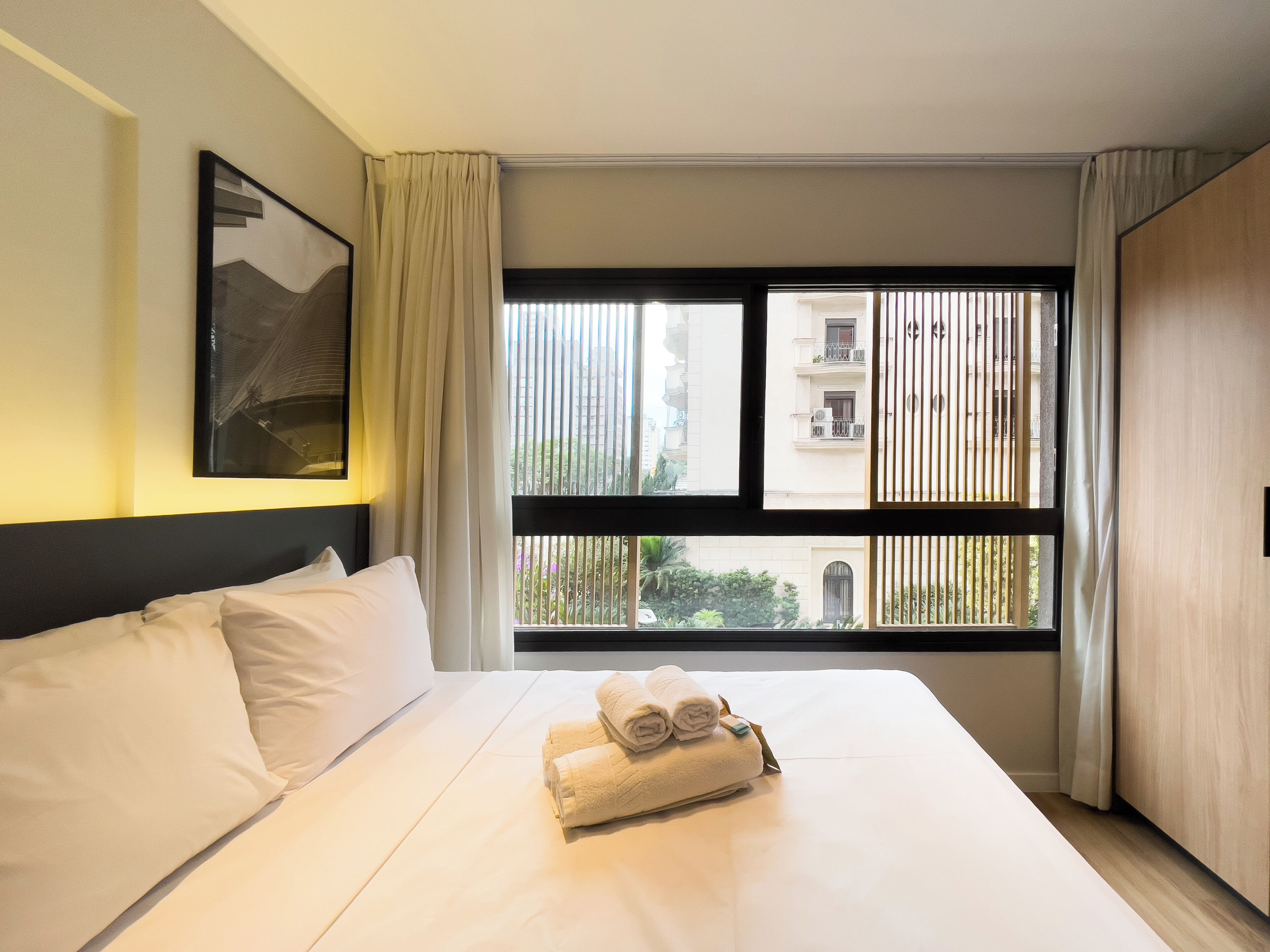 A hotel room with a bed, a window with blinds, a painting on the wall, and a towel on the bed.