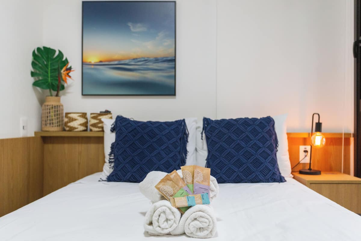 Et hotelværelse med en seng, to puder, et natbord med en lampe, et ophængt billede og en plante.