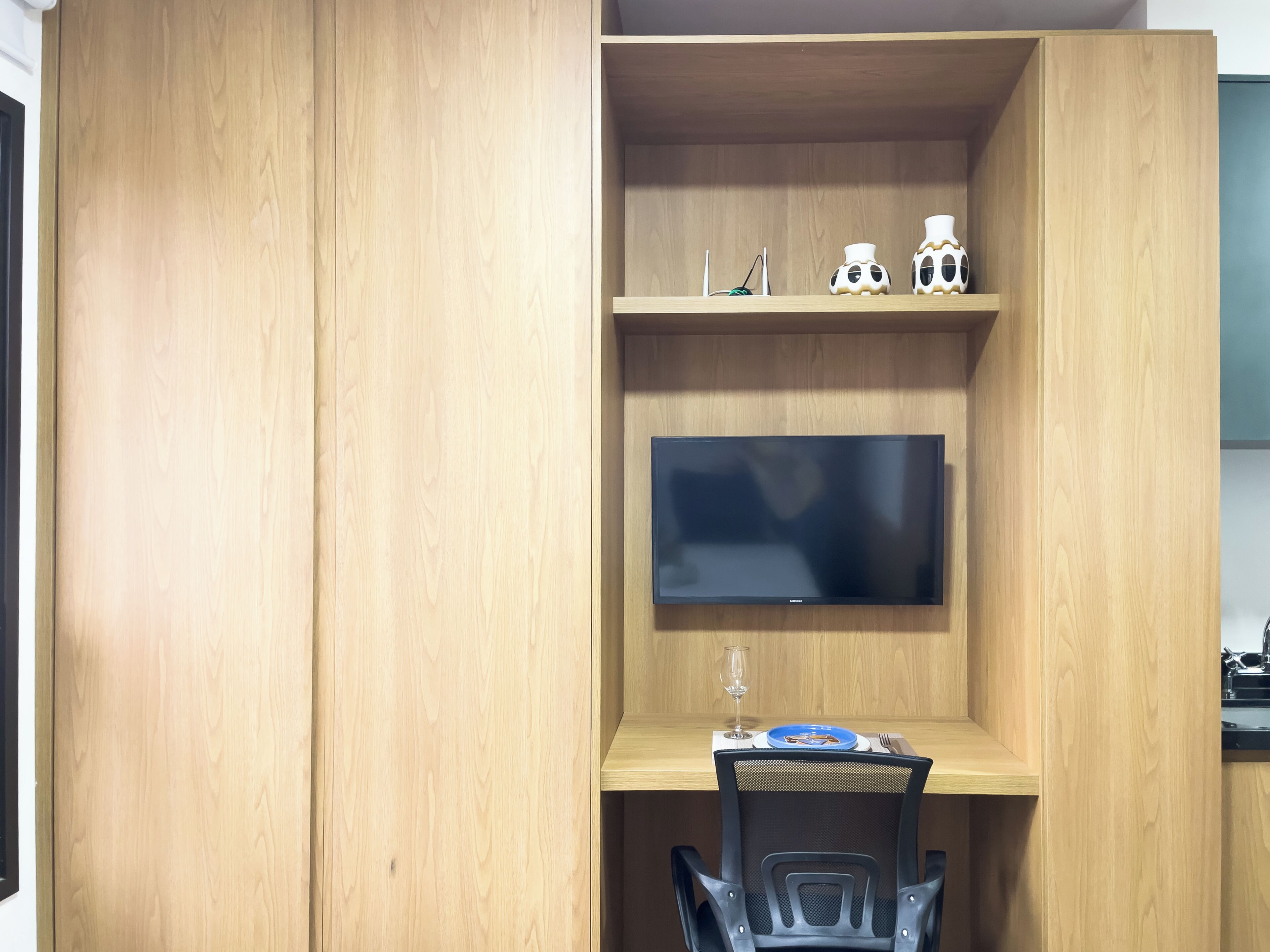 Ein Zimmer mit Holzschrank, flachbildfernseher an der Wand, Esstisch mit einem Stuhl und einem Weinglas.