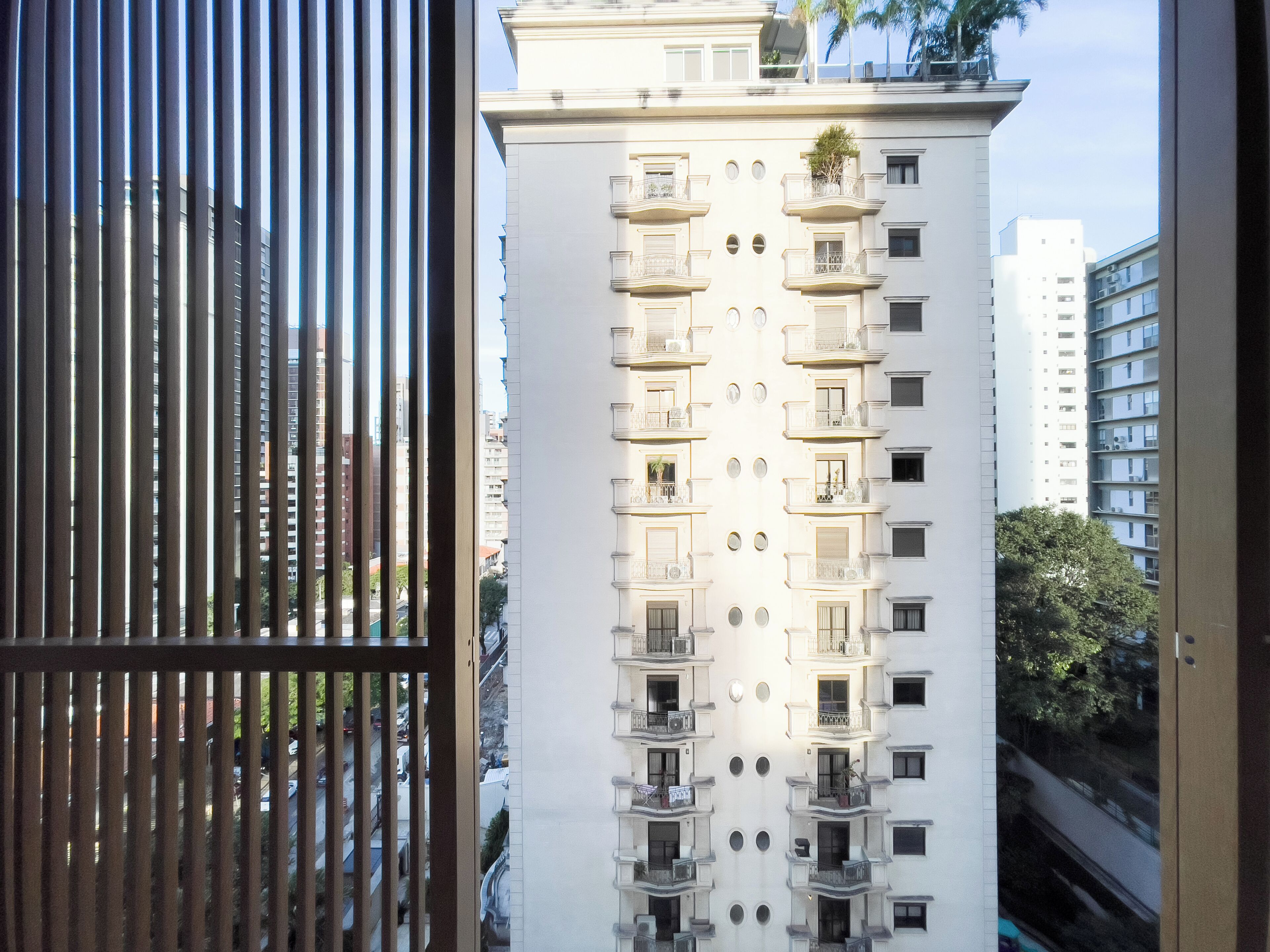 Un immeuble de grande hauteur, doté de nombreux balcons et fenêtres, vu depuis une fenêtre avec des stores verticaux.