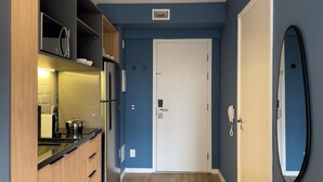 Ein schmaler Flur mit Küchenzeile auf der linken Seite, inklusive Kühlschrank, Herd und Holzschränken. Eine weiße Tür ist rechts zu sehen, und ein Spiegel hängt an der Wand.