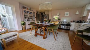 A spacious living room with a wooden dining table, green chairs, a chandelier, and a kitchen in the background.