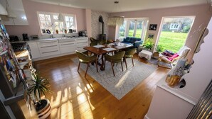 A cozy kitchen with wooden flooring, a dining table with chairs, a blue sofa, and a view of the garden through large windows.
