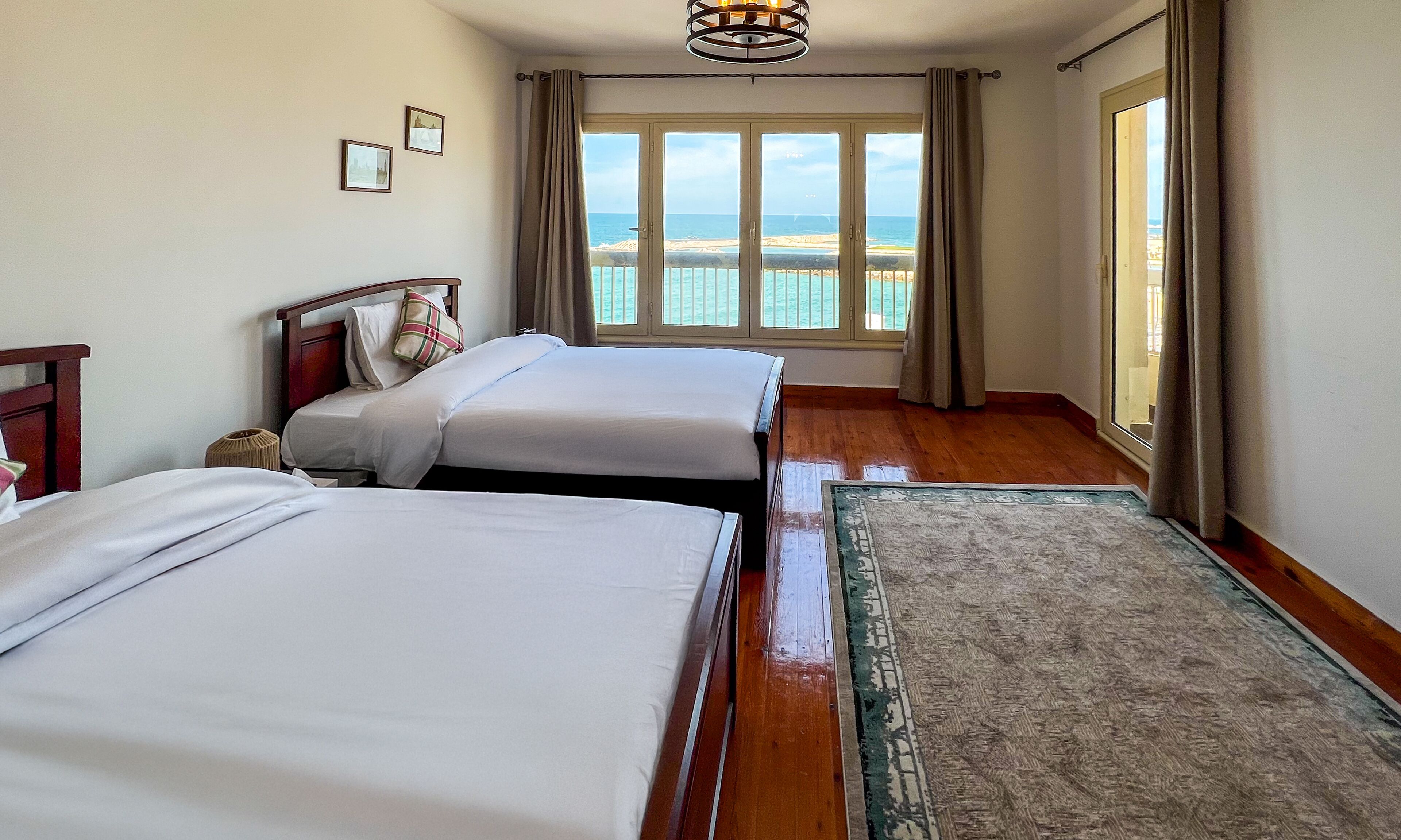 A hotel room with two beds, a wooden floor, a view of the sea, and a ceiling light fixture.