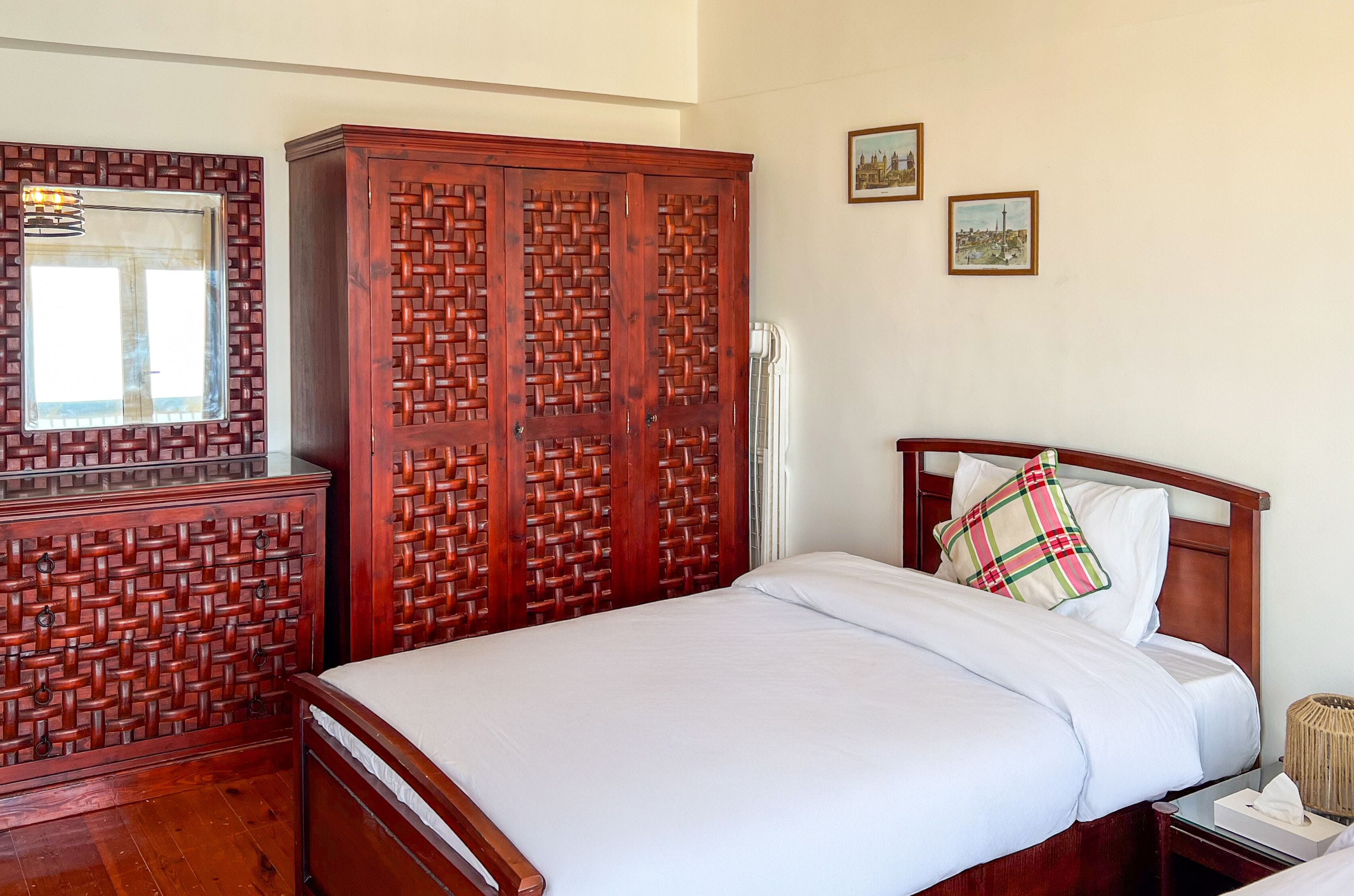 A neatly made bed with white linens, a wooden wardrobe with a woven pattern, a bedside table with a lamp, and two framed pictures on the wall.