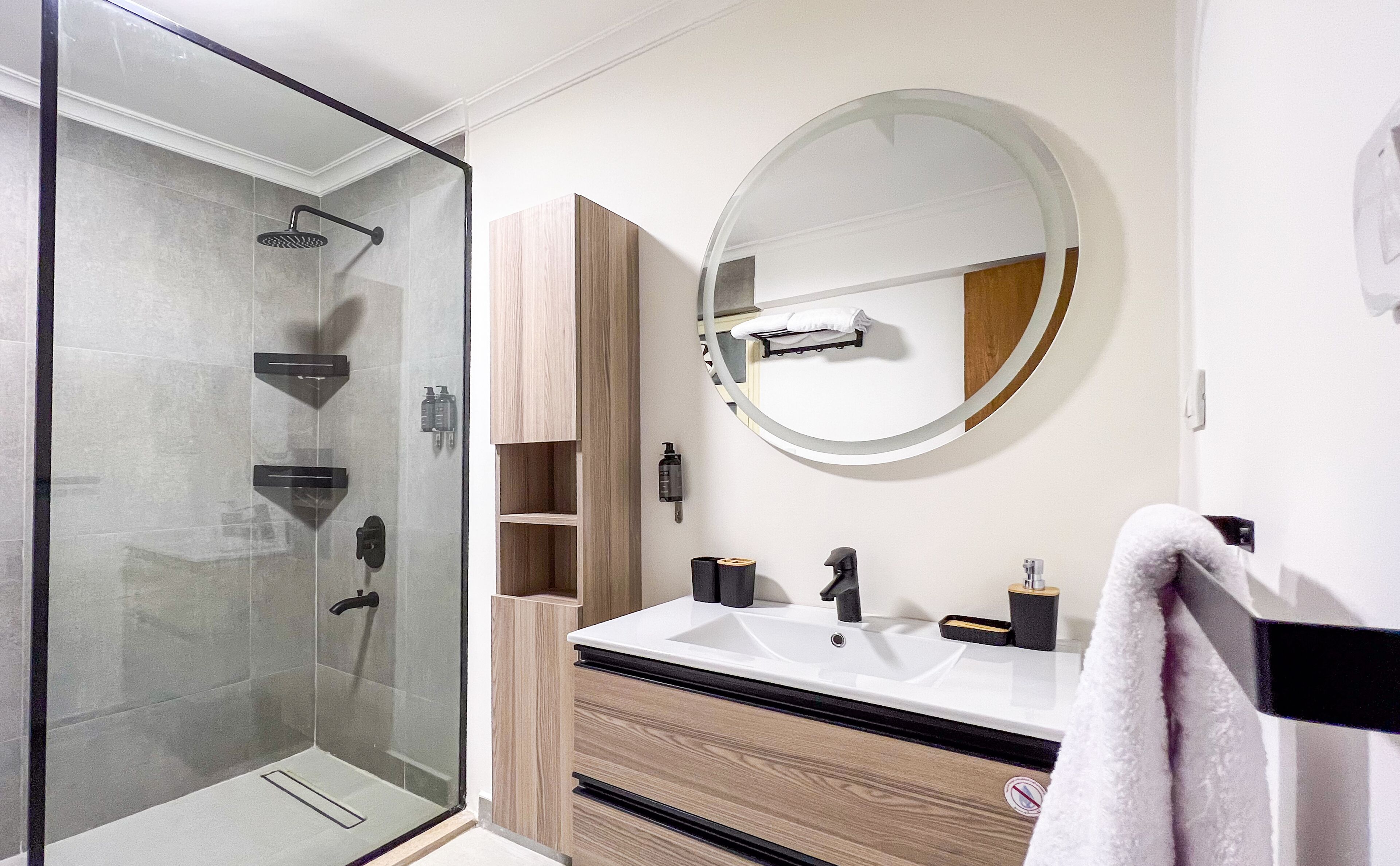 A modern bathroom with a glass-enclosed shower, a round mirror, and a wooden vanity with a white sink.