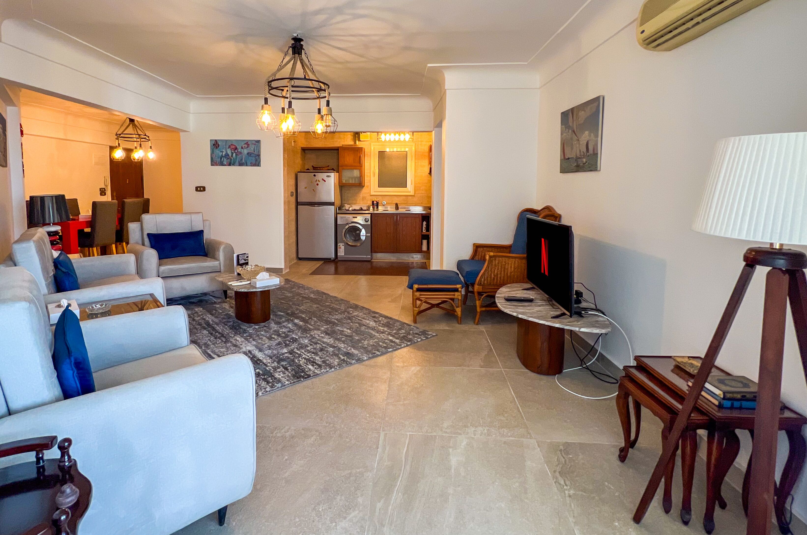 A living room with a sofa, armchair, and a TV. There is a kitchen area with a fridge, washing machine, and a dining table.