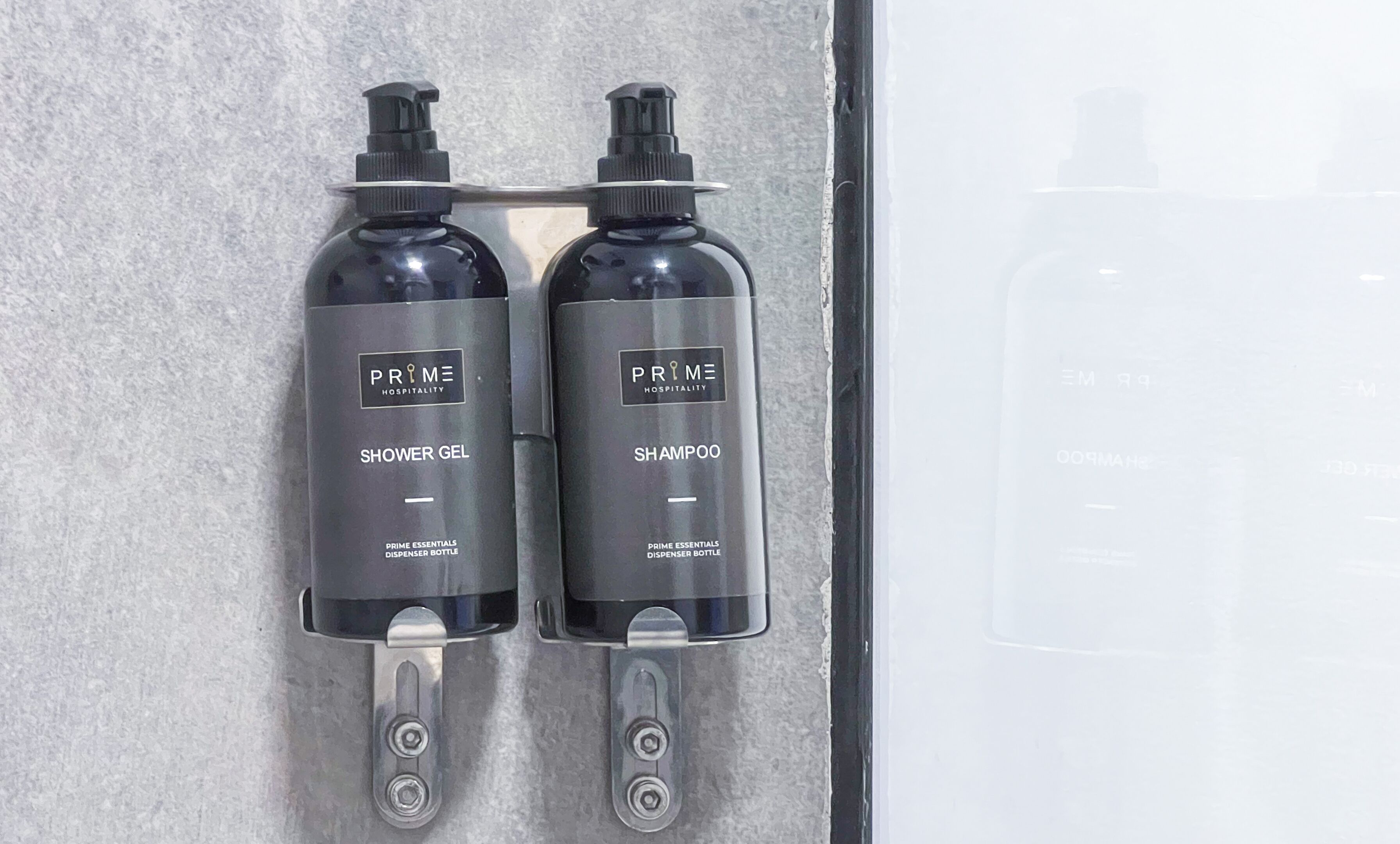 Dos botellas negras de Prime Hospitality, una etiquetada como 'Shower Gel' y la otra como 'Shampoo', sujetas a una superficie metálica.
