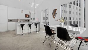 Una cocina moderna con una isla central, taburetes de bar, iluminación colgante y un amplio ventanal con vistas a un edificio.