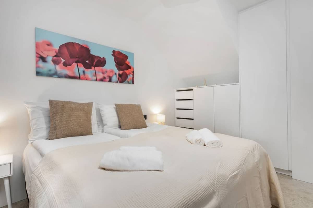 Een keurig opgemaakt slaapkamer met een bed, kussens, een nachtkastje en een kunstwerk aan de muur.