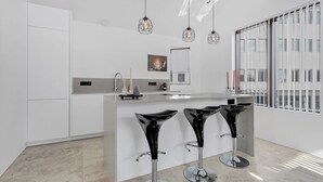 Una cocina moderna con armarios blancos, una isla central con taburetes de bar, iluminación colgante y un ventanal con persianas.
