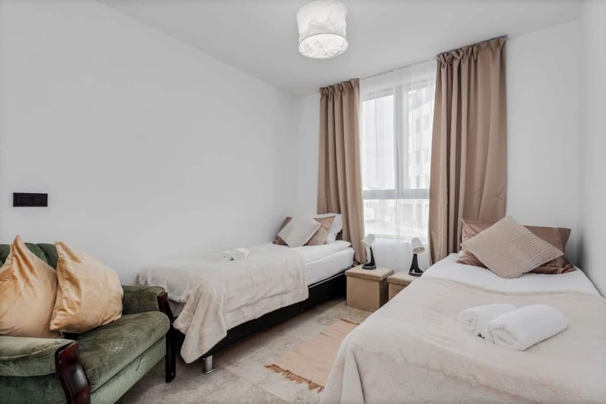 Een hotelkamer met twee bedden, een groene fauteuil, een nachtkastje met een lamp en een groot raam met gordijnen.