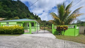 A green building with a gated entrance, a concrete driveway, and a palm tree. - Fully furnished 3BR home in Sylvania with Wi-Fi, AC, kitchen & serene views. (Sylvania)