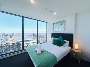 Une chambre d’hôtel avec une grande fenêtre, un lit avec une literie verte, une table de chevet avec une lampe et un tableau au mur.