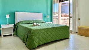 A bedroom with a green quilted bedspread, a white headboard, a nightstand with a card holder, and a view of the outdoors through a sliding glass door. - B&B Holiday (Torre Annunziata)