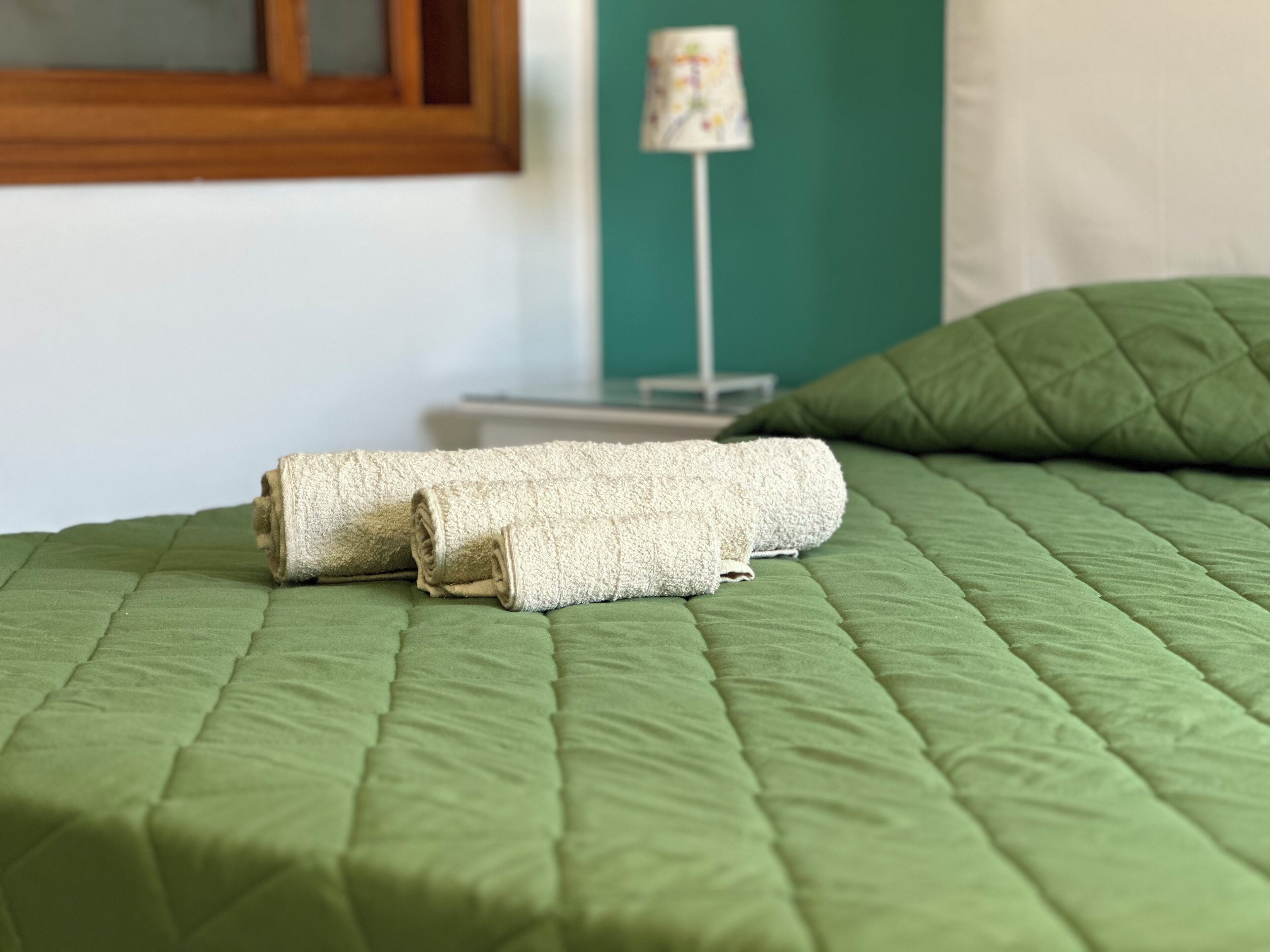 A neatly made bed with a green quilt, a rolled white towel, and a green pillow.