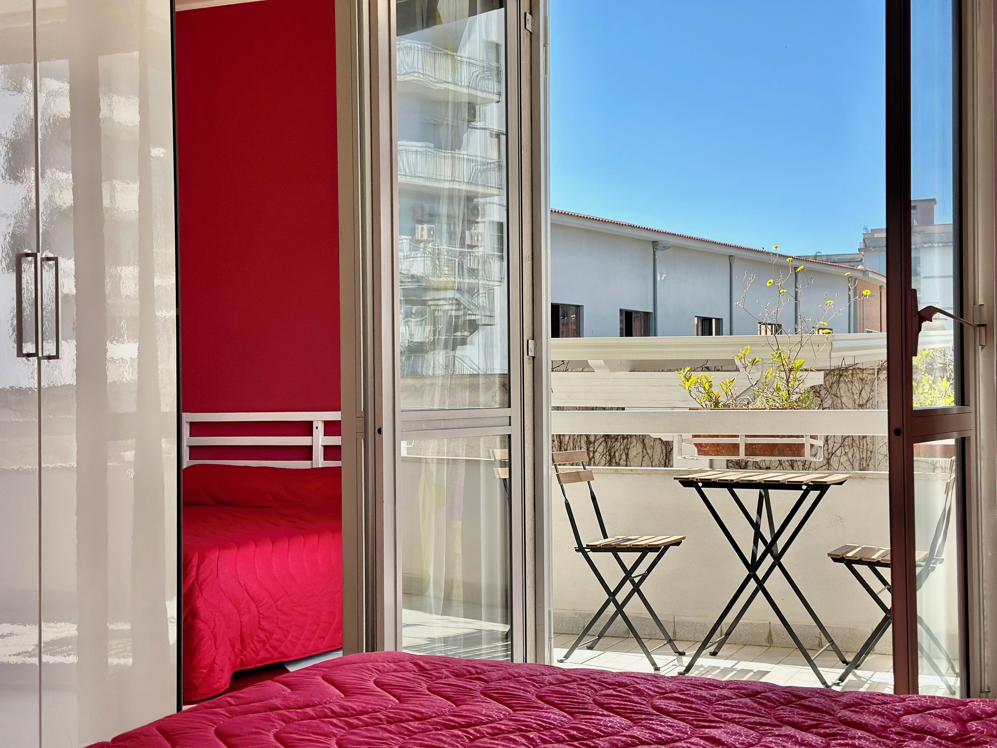 Une chambre avec une couvre-lit rouge et une porte-fenêtre coulissante donnant sur un balcon extérieur équipé d’une table et de chaises.