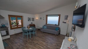 Ett kompakt bostadsutrymme med en matplats, ett litet kök och en soffa som är placerad framför ett stort fönster med utsikt över havet.
