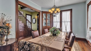 Une salle à manger avec une longue table, des chaises en bois, un lustre et un escalier menant à une autre pièce.