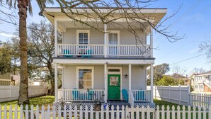 Une maison à deux étages avec une porte d’entrée verte, une clôture blanche en bois et un balcon au deuxième étage.