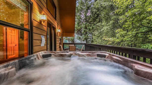 A hot tub on a wooden deck with a view of a forest. - Count Your Blessings 3 Bedroom Home (Pigeon Forge)