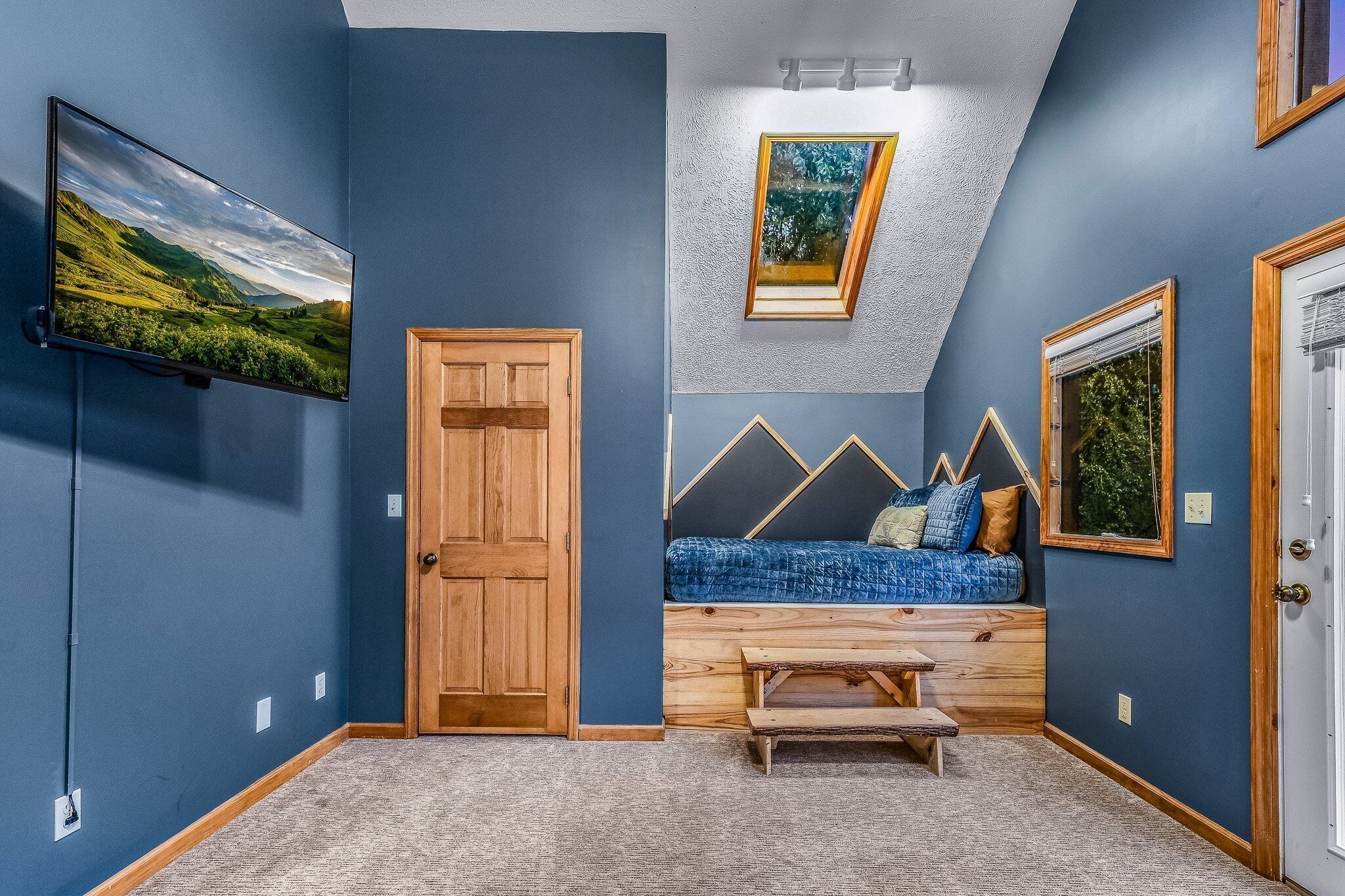 A room with a sloped ceiling, a mounted TV, a wooden door, a built-in bench with a blue cushion, and framed pictures on the walls.