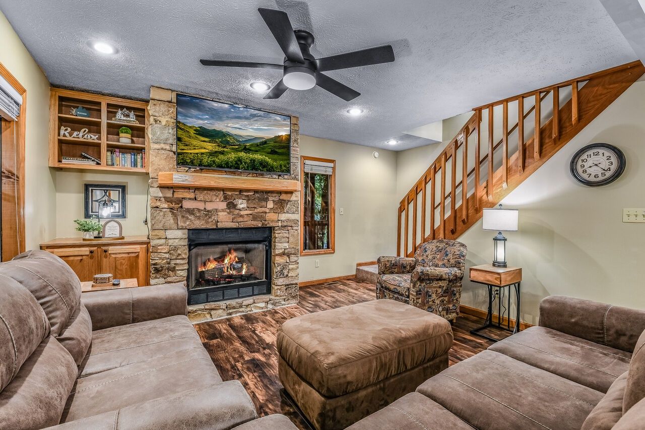 A living room with a fireplace, a ceiling fan, a staircase, and a bookshelf.