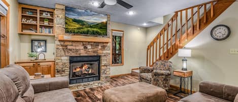 A living room with a fireplace, a ceiling fan, a staircase, and a bookshelf.