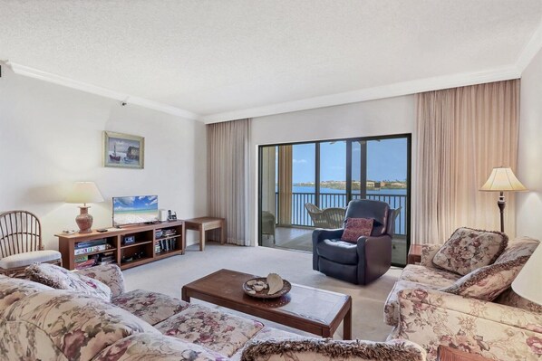 A spacious living room with a sofa set, a coffee table, a TV, and a bookshelf. There is a balcony with a view of the water. - 2/2 Bay Front Condo, Pool, Private Beach Access (Longboat Key)
