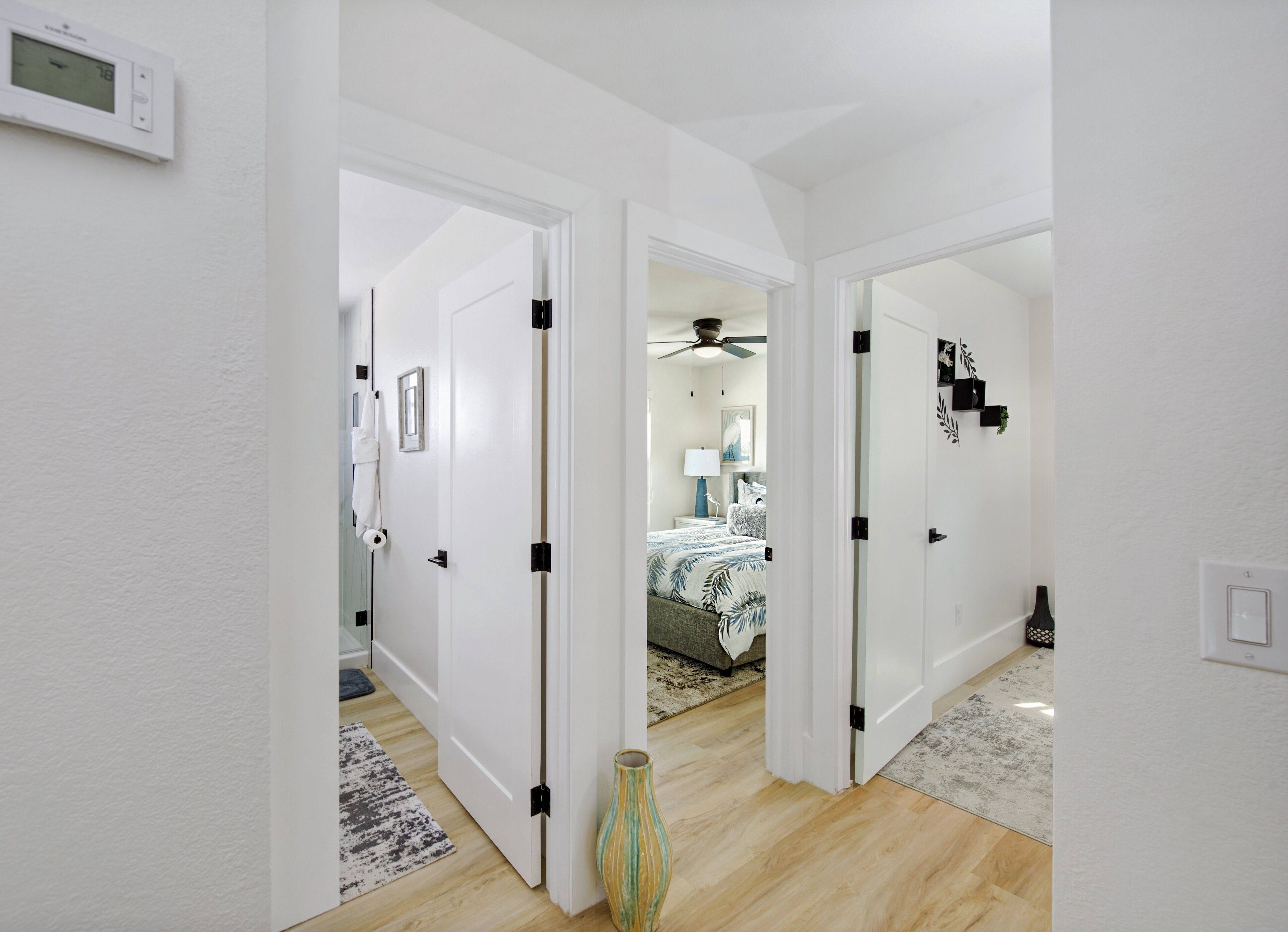A hallway with white walls, wooden flooring, and a ceiling fan. There are two doors leading to bedrooms, one with a bed and the other with a nightstand and lamp.