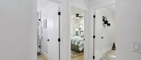 A hallway with white walls, wooden flooring, and a ceiling fan. There are two doors leading to bedrooms, one with a bed and the other with a nightstand and lamp.