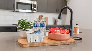 Uma bancada de cozinha com uma tábua de madeira que suporta uma planta, duas garrafas de água, um recipiente com lanches e uma tigela de batatas chips.