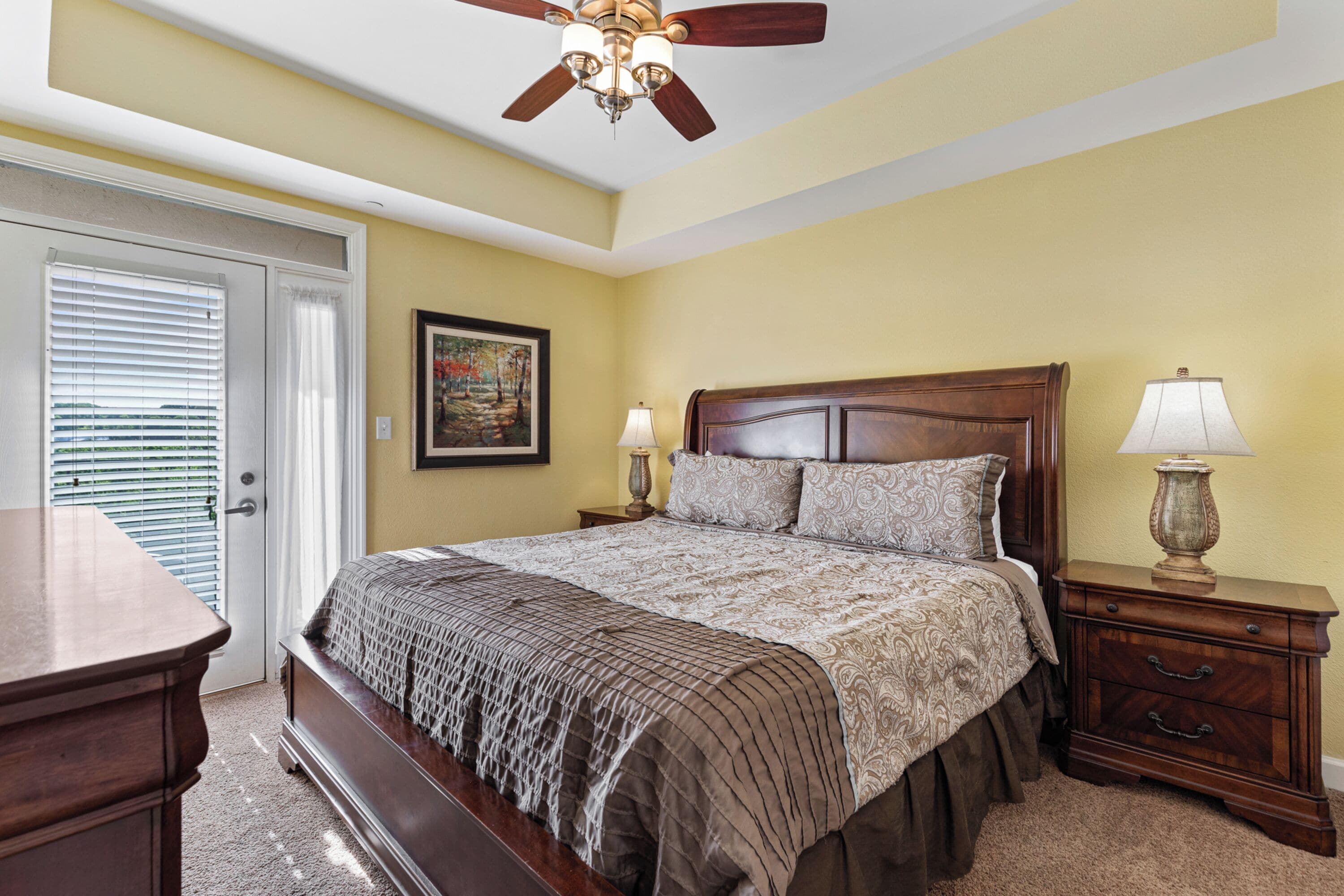A bedroom with a bed, bedside tables, a ceiling fan, and a door with blinds.