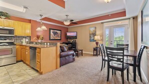 A modern kitchen with wooden cabinets, a built-in oven, and a dishwasher. There is a dining area with a table and chairs, and a living room with a sofa and a TV.