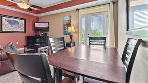 A dining area with a wooden table, black chairs, a ceiling fan, a television, and a painting on the wall.