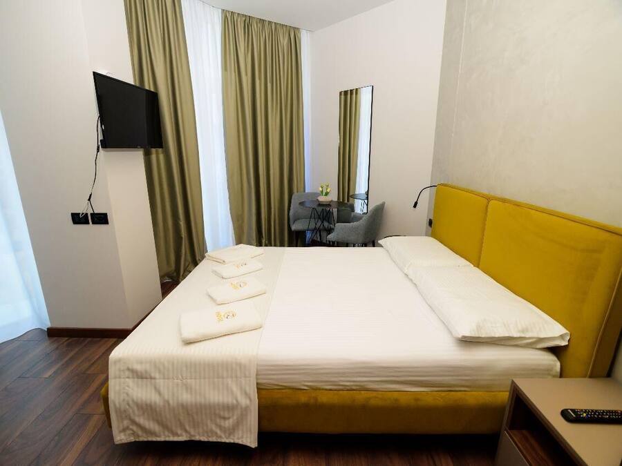 A hotel room with a bed, a TV mounted on the wall, a desk with a chair, and a window with curtains.