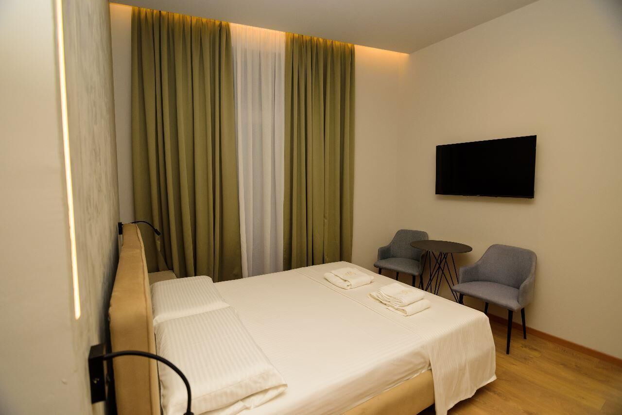 A hotel room with a bed, a TV mounted on the wall, and two chairs near a small table.