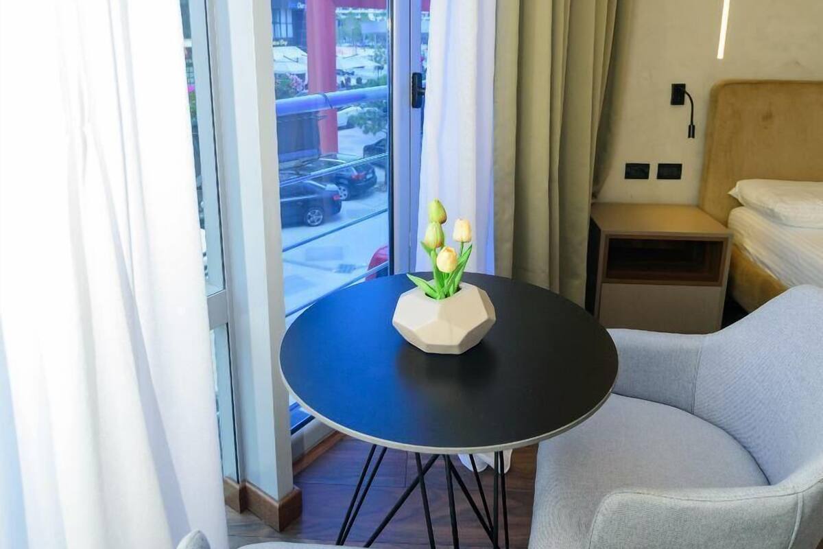 A hotel room with a balcony, a round table with a vase of flowers, two grey armchairs, and a bed with a wooden headboard.
