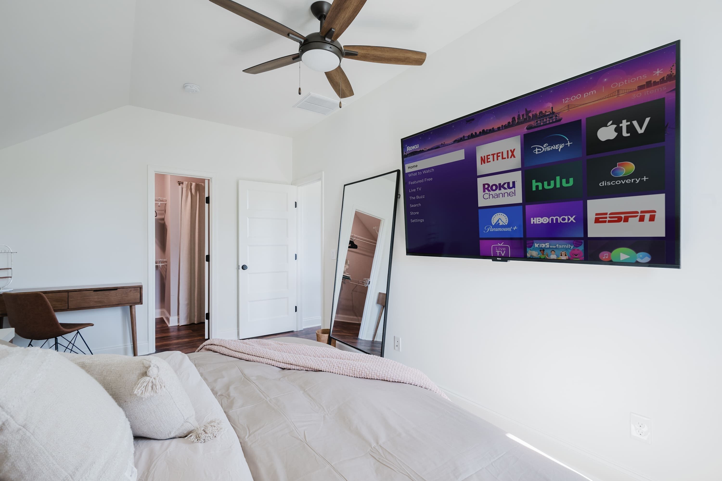 A bedroom with a bed, a desk, a ceiling fan, a TV wall-mounted with various streaming service logos, and a door leading to another room.