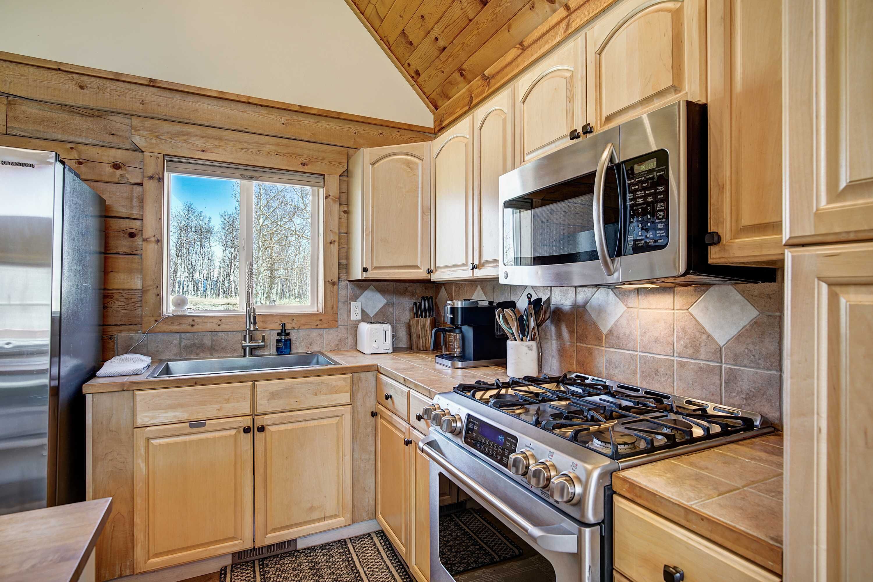A modern kitchen with wooden cabinets, stainless steel appliances, and a large window offering a view of trees.