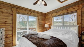 Une chambre dans une cabane en bois, avec un lit, une commode, une lampe et de grandes fenêtres.