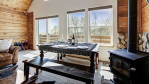 Une pièce chaleureuse avec un plafond en bois, une cheminée en pierre et de grandes fenêtres donnant sur la forêt.