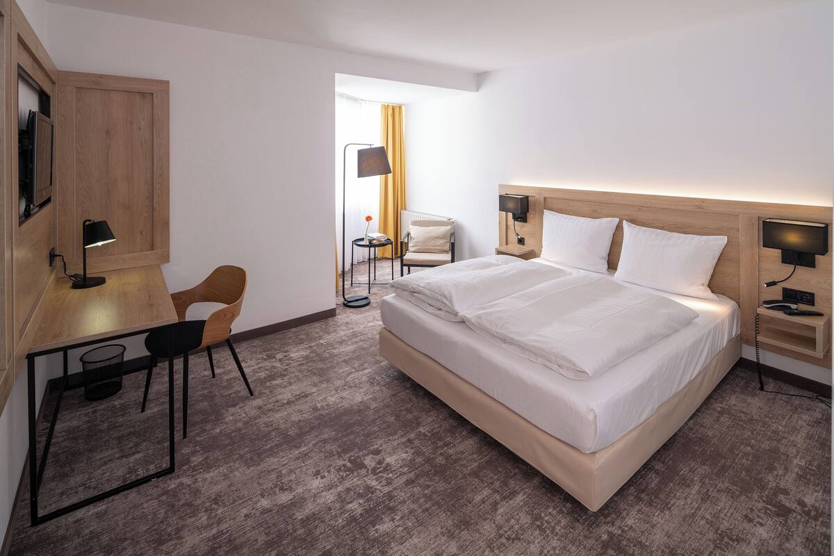 Ein Hotelzimmer mit Bett, Schreibtisch, Stuhl und einem kleinen Tisch mit Lampe.