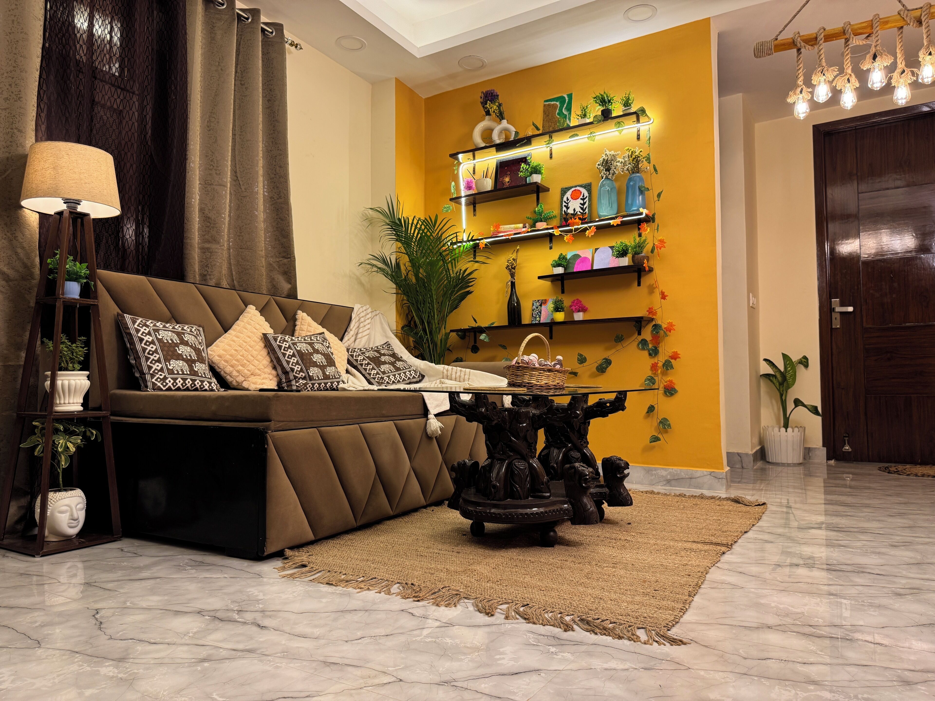A living room with a brown sofa, patterned cushions, a coffee table, and a wall-mounted shelf with plants and decorative items.