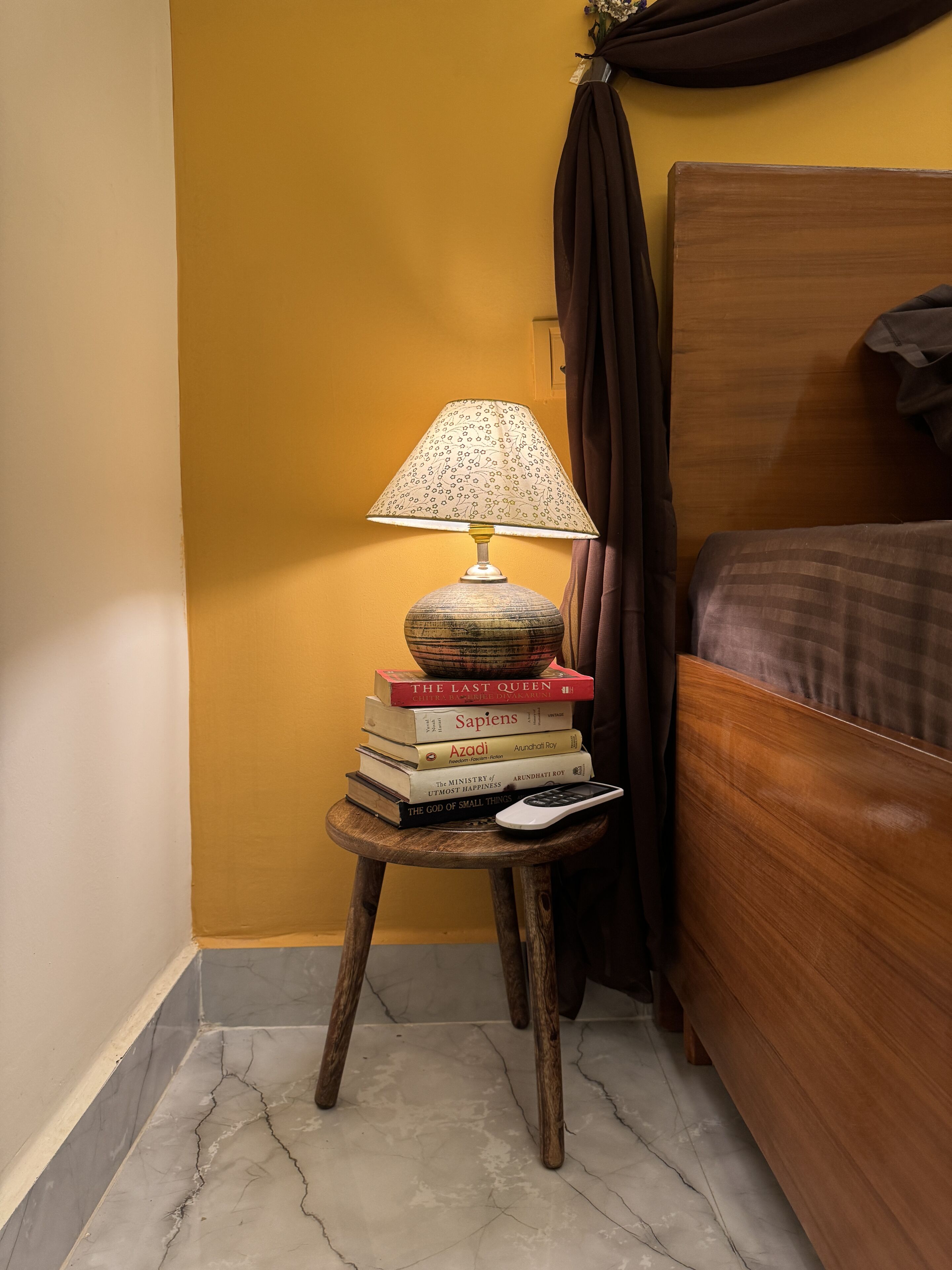 Een houten nachtkastje met een lamp, een stapel boeken en een afstandsbediening.