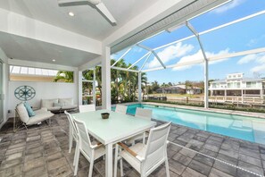 Un espace piscine extérieur moderne, doté d’une table et de chaises pour les repas, d’un ventilateur de plafond et offrant une vue sur un quartier résidentiel.