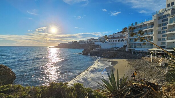 A coastal hotel with a beach view, a rocky shoreline, and a clear sky.