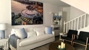 A living room with a white sofa, a round coffee table with candles, a large painting of a coastal scene, and a staircase.